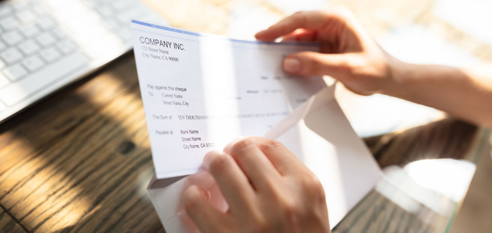 Close-up Of A Businessperson's Hand Opening Envelope With Paycheck Over Wooden Desk